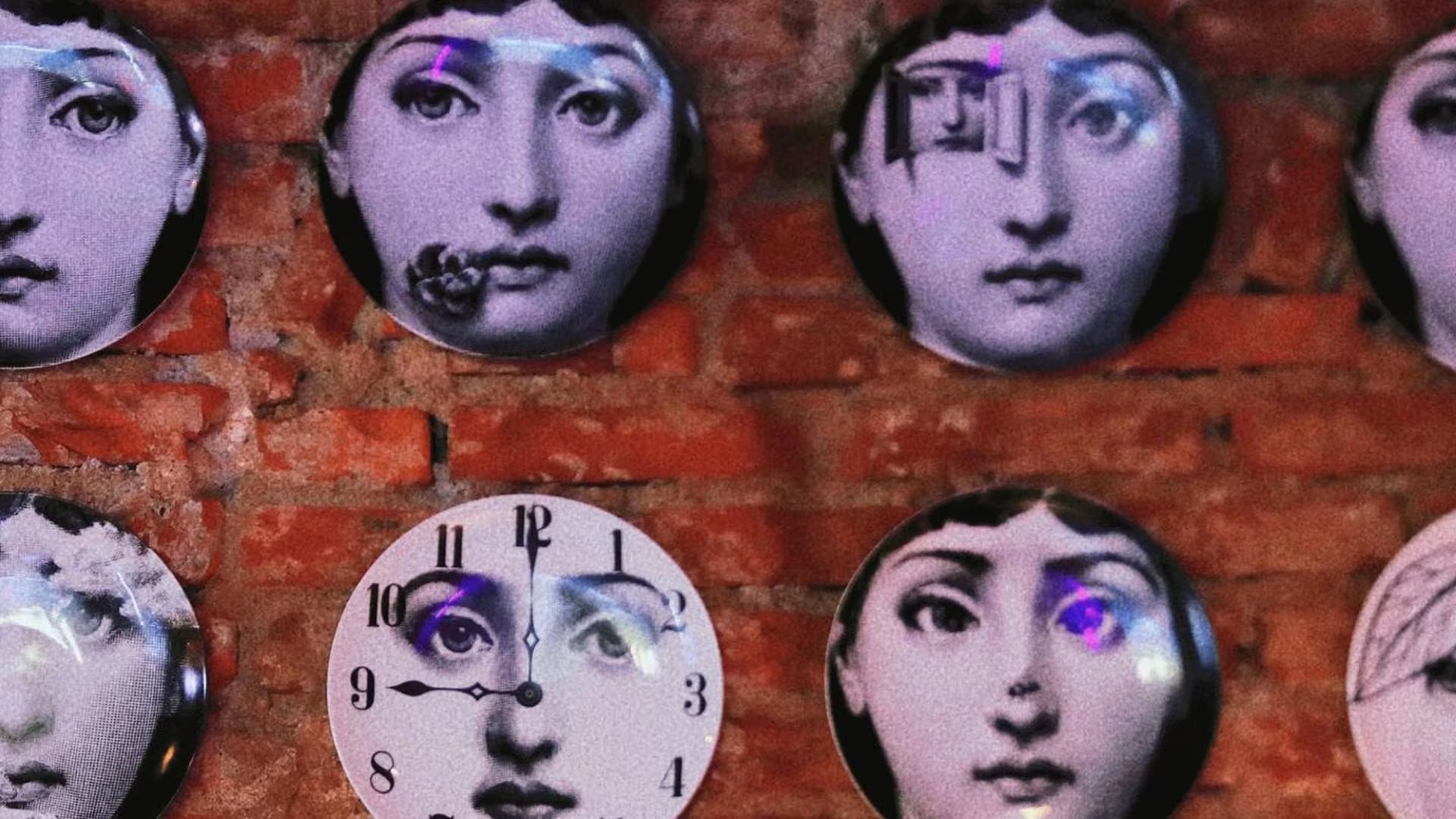 Parete di mattoni con piatti decorativi appesi, ciascuno con un volto femminile stampato in bianco e nero, accesi da una luce viola.