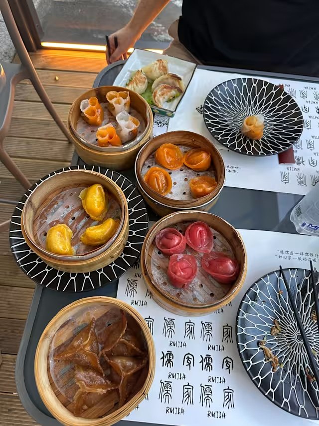 Tavola apparecchiata con sei cestelli di bambù di dim sum colorati — rosa, arancioni, gialli e ambrati — sulle tovagliette con il logo 儒家.