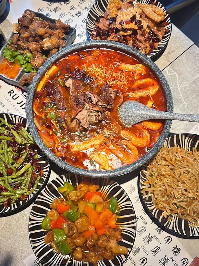 Tavolata condivisa: una grande zuppa piccante al centro, circondata da fagiolini al peperoncino, pollo in agrodolce, noodles saltati e frittura croccante.