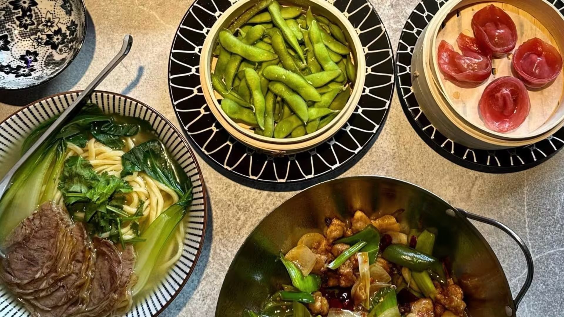 Vista dall'alto di una tavola: ramen di manzo con bok choy, edamame nel cestello, ravioli rossi al vapore e un padellino di pollo saltato.
